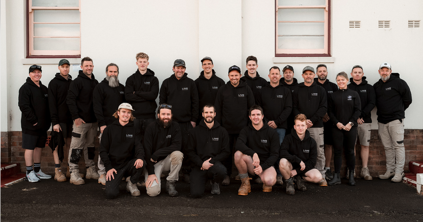 Group photo of Kiama & Berry Builder Lime Building Group wearing black team hoodies standing against white brick wall.